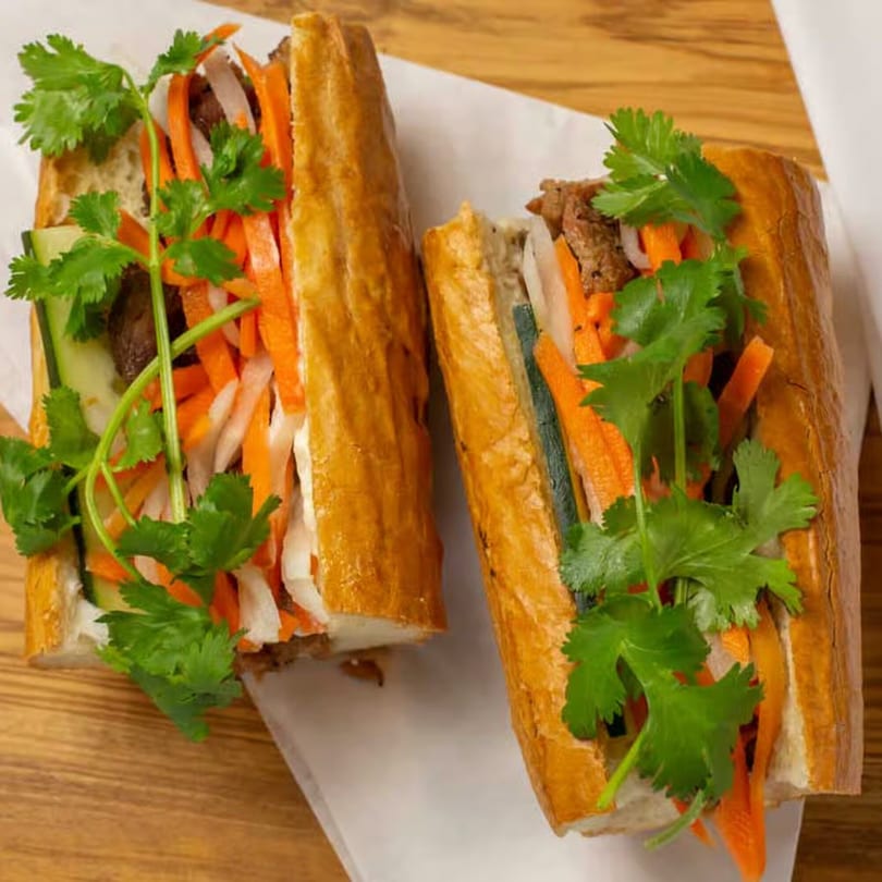 Two Vietnamese banh mi sandwiches, cut in half, filled with meat, pickled carrots, cucumbers, and cilantro.