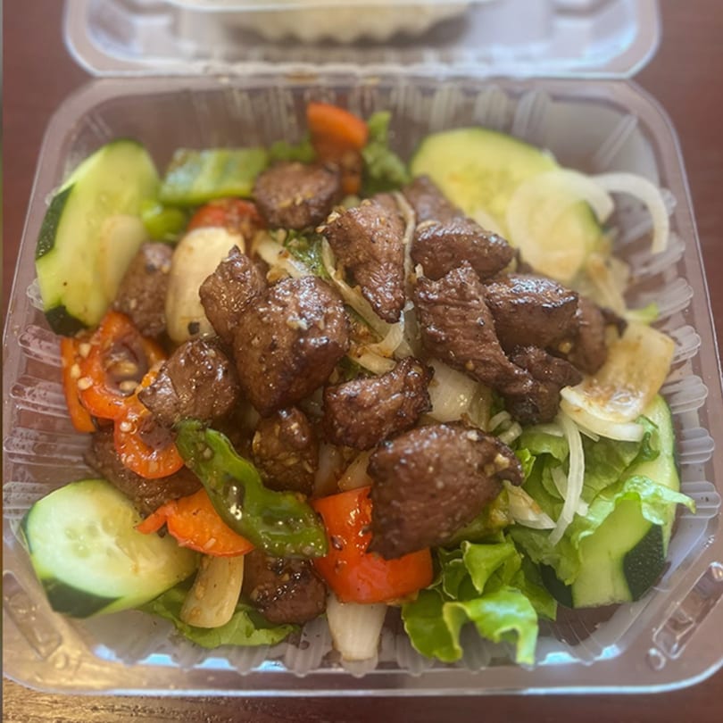 Vietnamese shaken beef salad with lettuce, cucumbers, onions, and bell peppers in a plastic container.