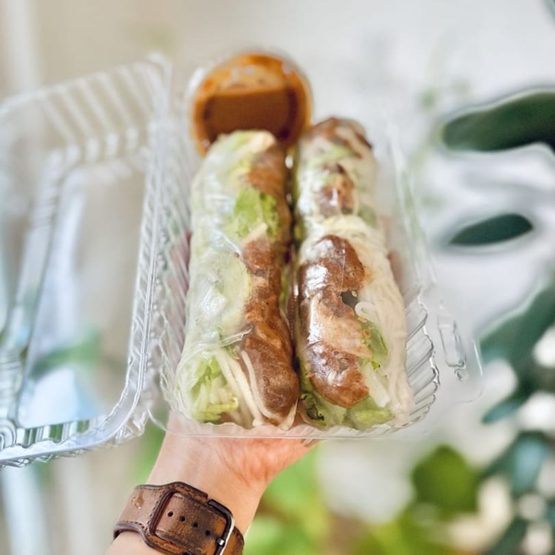 Two Vietnamese spring rolls and dipping sauce in a plastic takeout container, held by a hand.